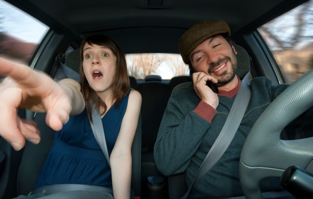 A scared young woman in the passenger seat trying to alert the driver talking on his phone that he’s about it get in an accident.