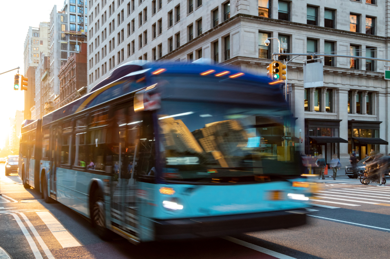 A blurred image of a blue bus driving down the street with sunlight shining between the buildings in the background