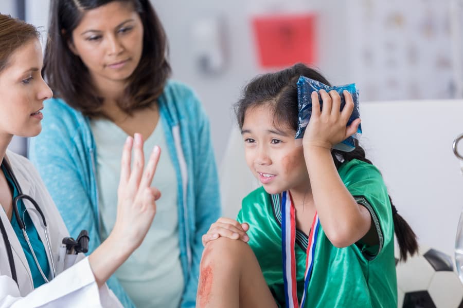 Female ER doctor holds up three fingers while talking with young injured female soccer player holding an ice pack to her head.