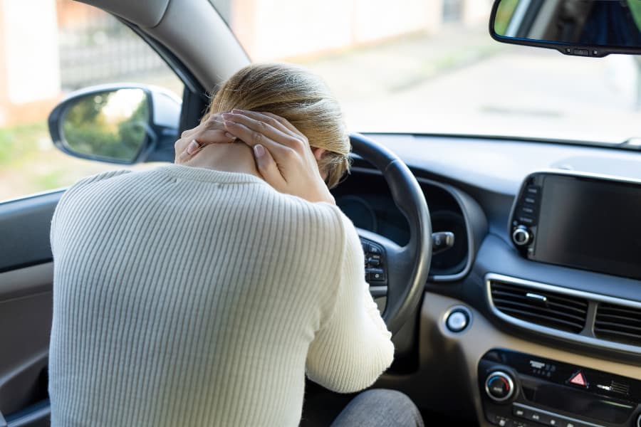 Injured woman holding her neck and leaning her forehead on the steering wheel in a car.