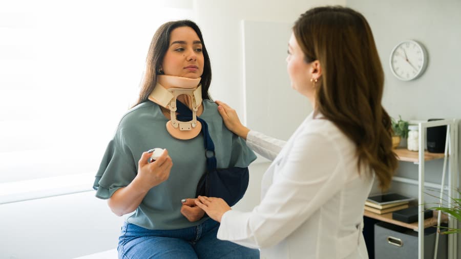 A young woman with a cervical collar and arm sling with a female doctor examining and comforting her.