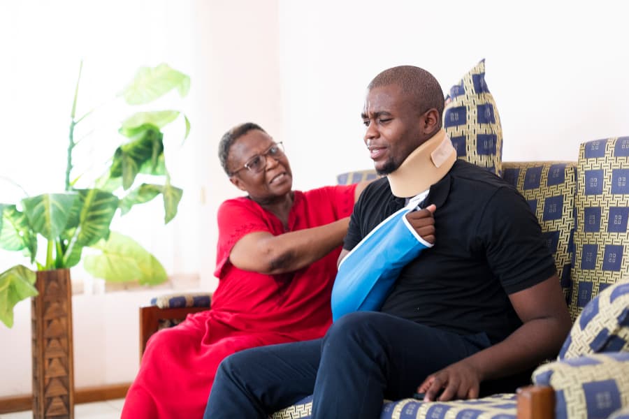 An older woman adjusting the cushions on a couch for her injured son wearing a cervical collar and arm sling.