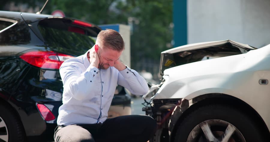 A man holding his neck while crouching in front of two wrecked cars after an accident.