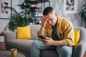 A stressed out man sitting on a couch holding his head and frowning at his phone.