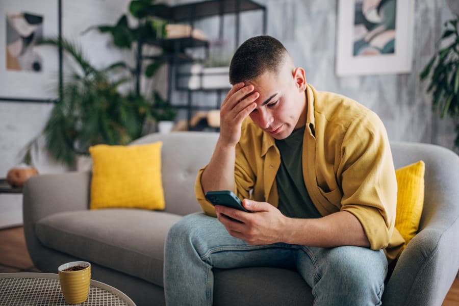 A stressed out man sitting on a couch holding his head and frowning at his phone.
